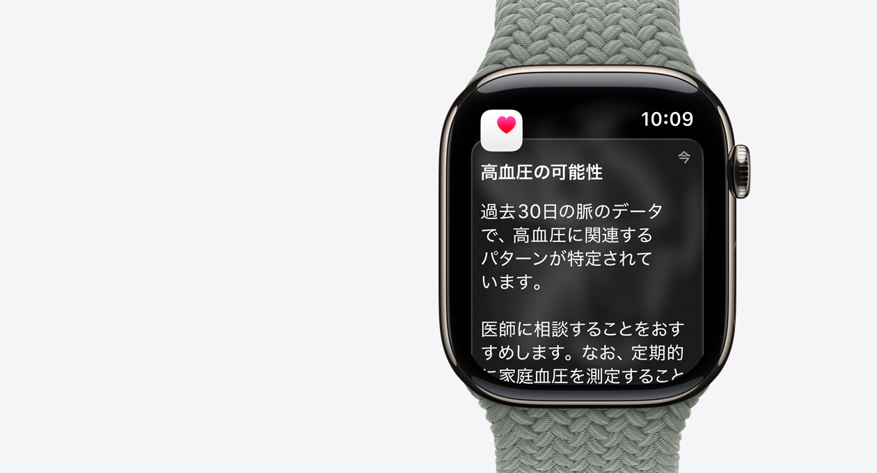 ナチュラルチタニウムケースのApple Watch Series 11。高血圧の兆候を示す通知と、ピンクのハートマークが入った白いボックスが表示されている。右側面の外観にはDigital Crownがあり、グリーングレイのブレイデッドソロループが付いている
