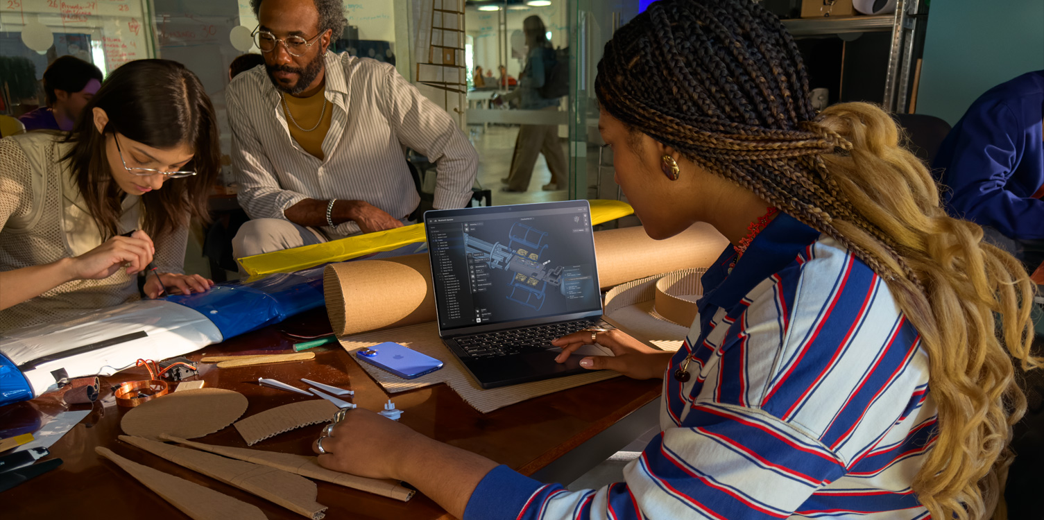 A group of students sitting at a table working on a technical project. One has an open MacBook in front of her, displaying the Shapr 3D app.