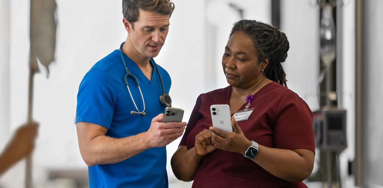 A male provider in blue scrubs looks at his iPhone and shows something on it to a female provider in red scrubs, who also has an iPhone.