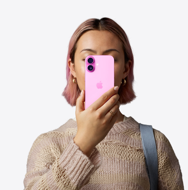 A person holding iPhone16 in pink finish in front of her face showing back exterior, dual camera system on left top corner.