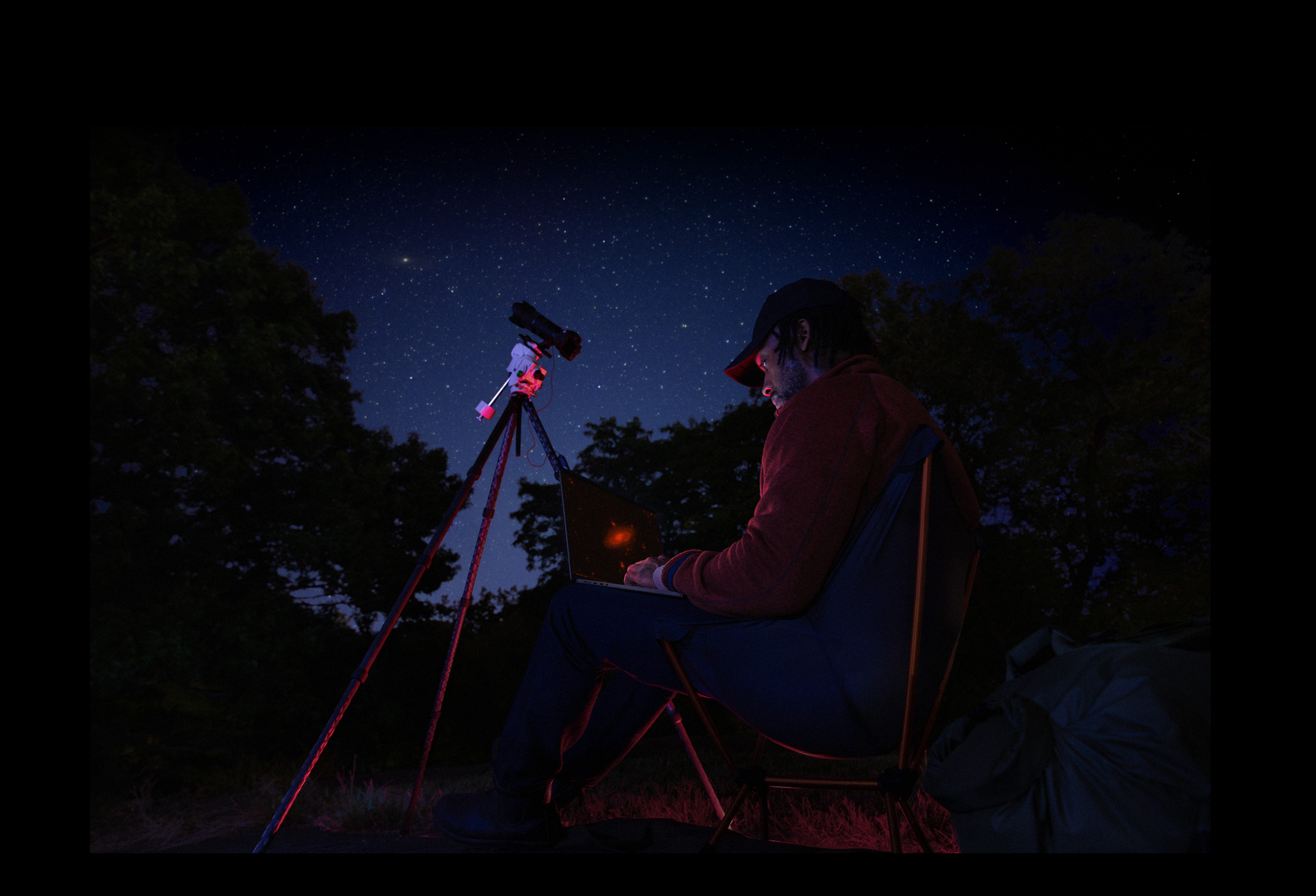 A person outside at night using a MacBook Pro on their lap, connected to a device on a tripod pointed toward the starry sky