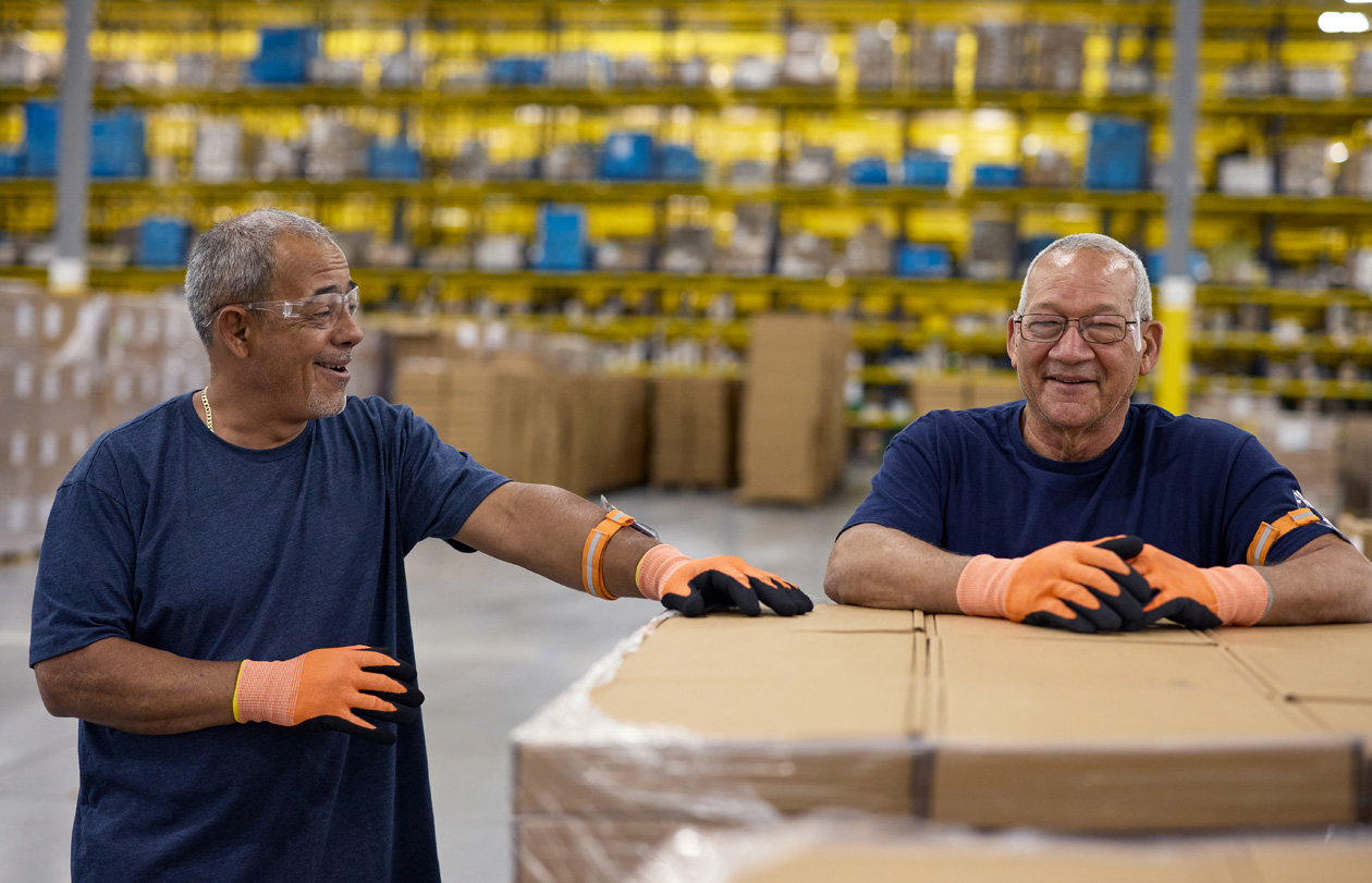 Employees wearing safety glasses and gloves stand by a pallet of boxes in a warehouse