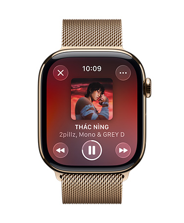 Apple Watch màu Titan Gold với dây quấn Milan, mặt đồng hồ hiển thị Apple Music đang phát, nút điều khiển dạng Liquid Glass trong mờ